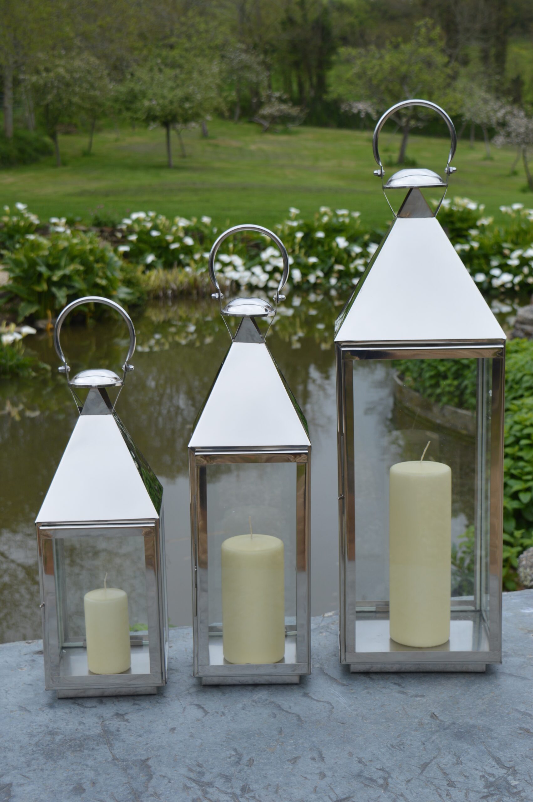 Set 3 stainless steel candle lanterns by pond surrounded by flowers