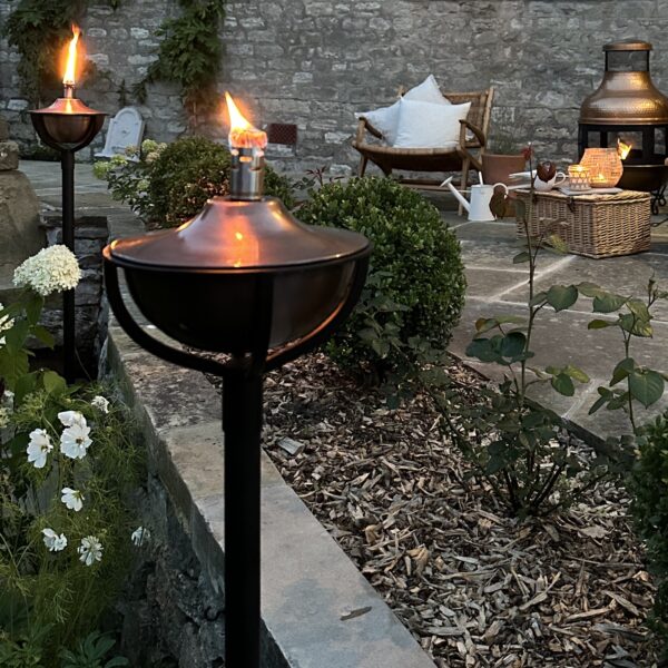 Aged copper torches on poles in garden setting, with garden furniture in background