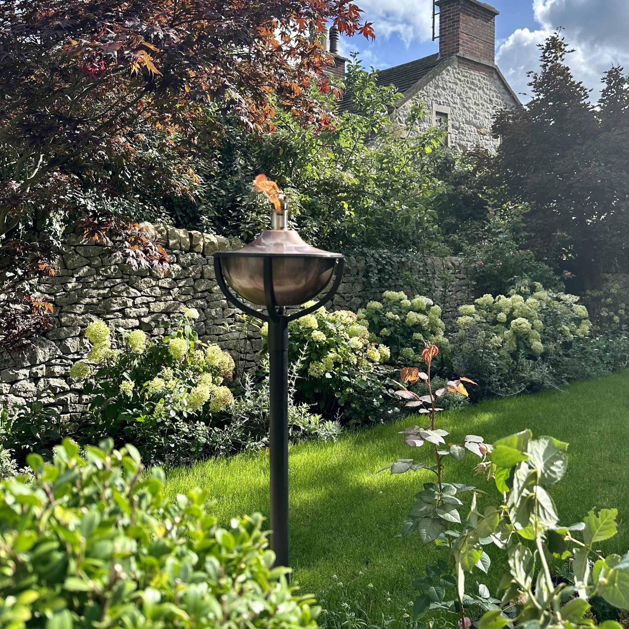 Aged copper Classica oil torch on black pole in sunny garden setting, in front of stone wall on lawn