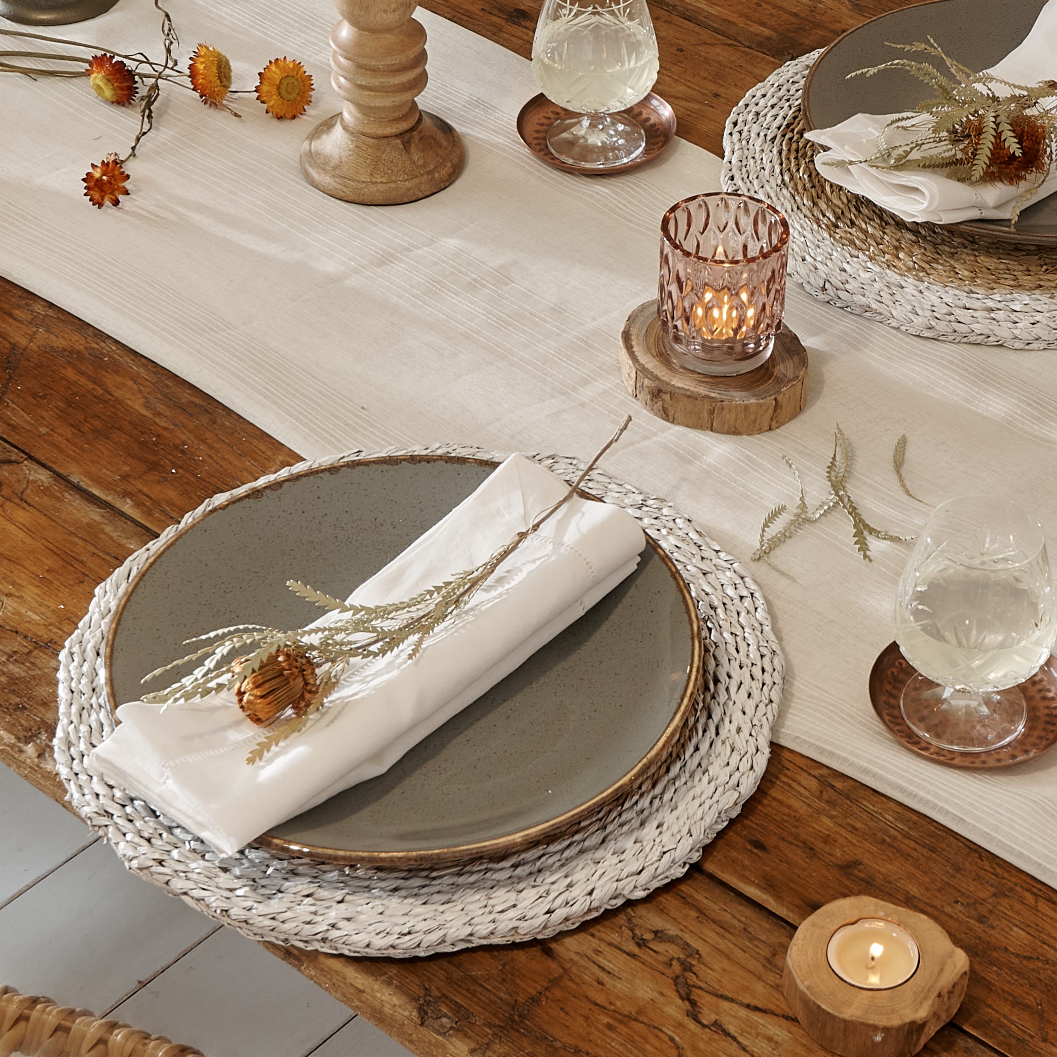 White wicker placemat on wooden table with cotton runner, with wooden and glass candlesticks, wooden and copper coasters and dried flowers