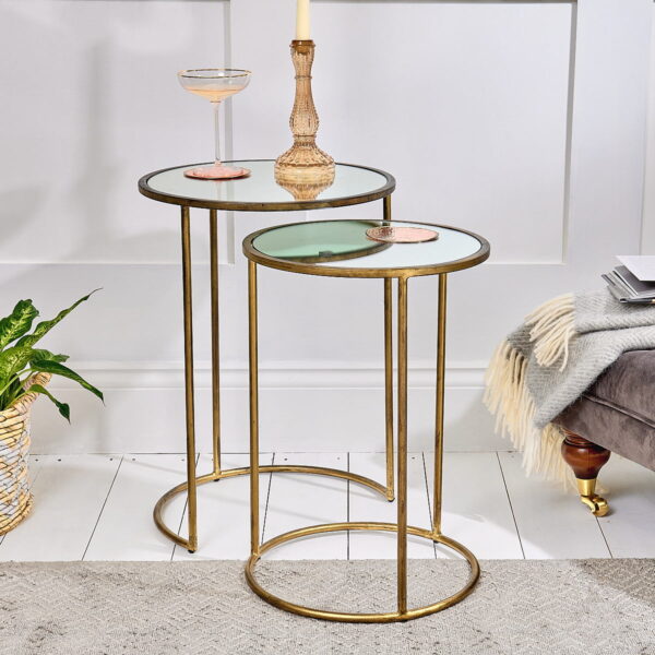 Brass and glass nest of tables with candle and drink on top next to a plant and lounge chair.