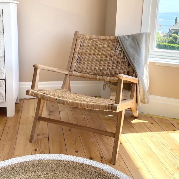 Teak and rattan armchair on wooden floor in bedroom setting by window, with wicker rug and white sideboard