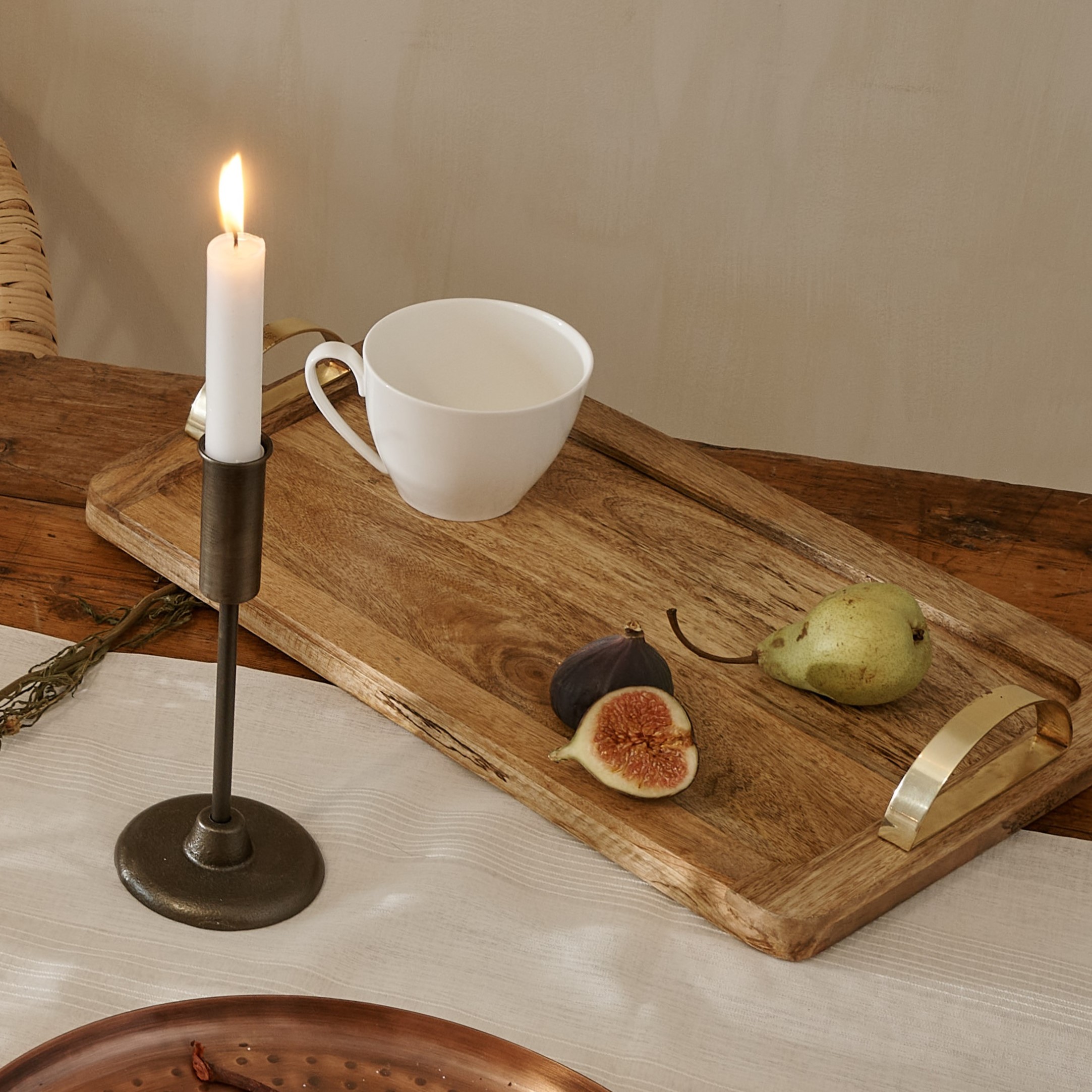 Wooden serving tray with copper handles on wooden table, with white mug and fruit, with antique copper candlestick with white candle