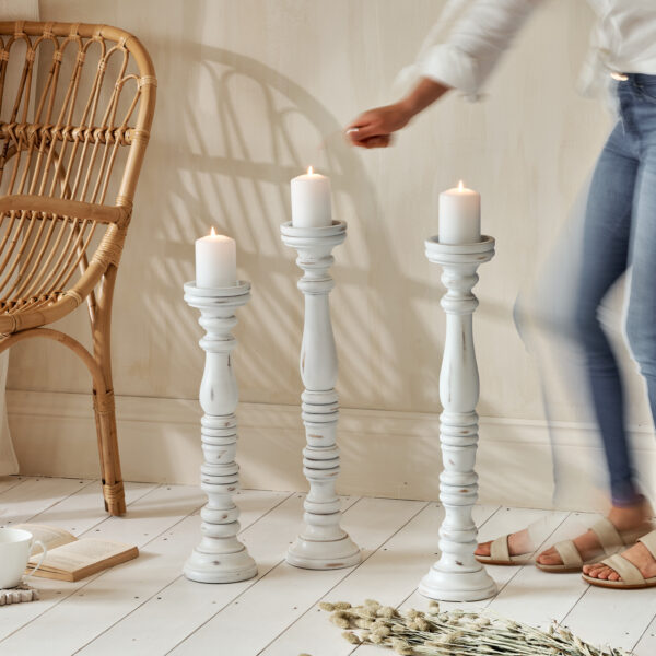 Tall wooden candle holders on white wooden floor, with bamboo chair. Woman lighting candles