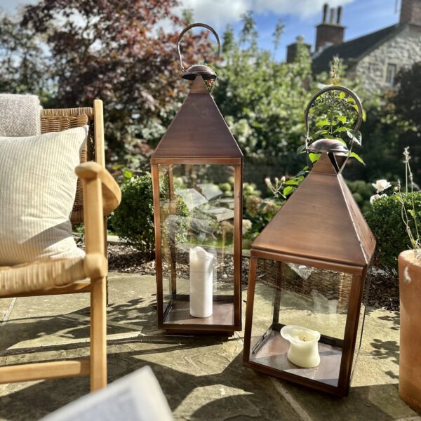 Two aged copper lanterns outdoors in garden setting, next to rattan chair on sunny patio