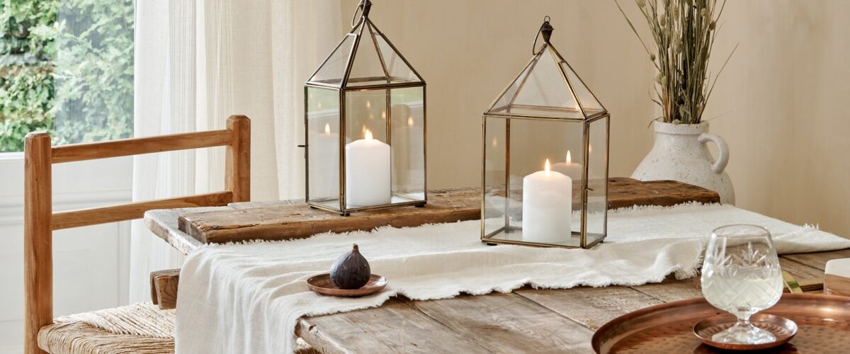 Small glass and brass candle lanterns on cotton runner on wooden table, with copper tray and coaster, with wicker dining chair