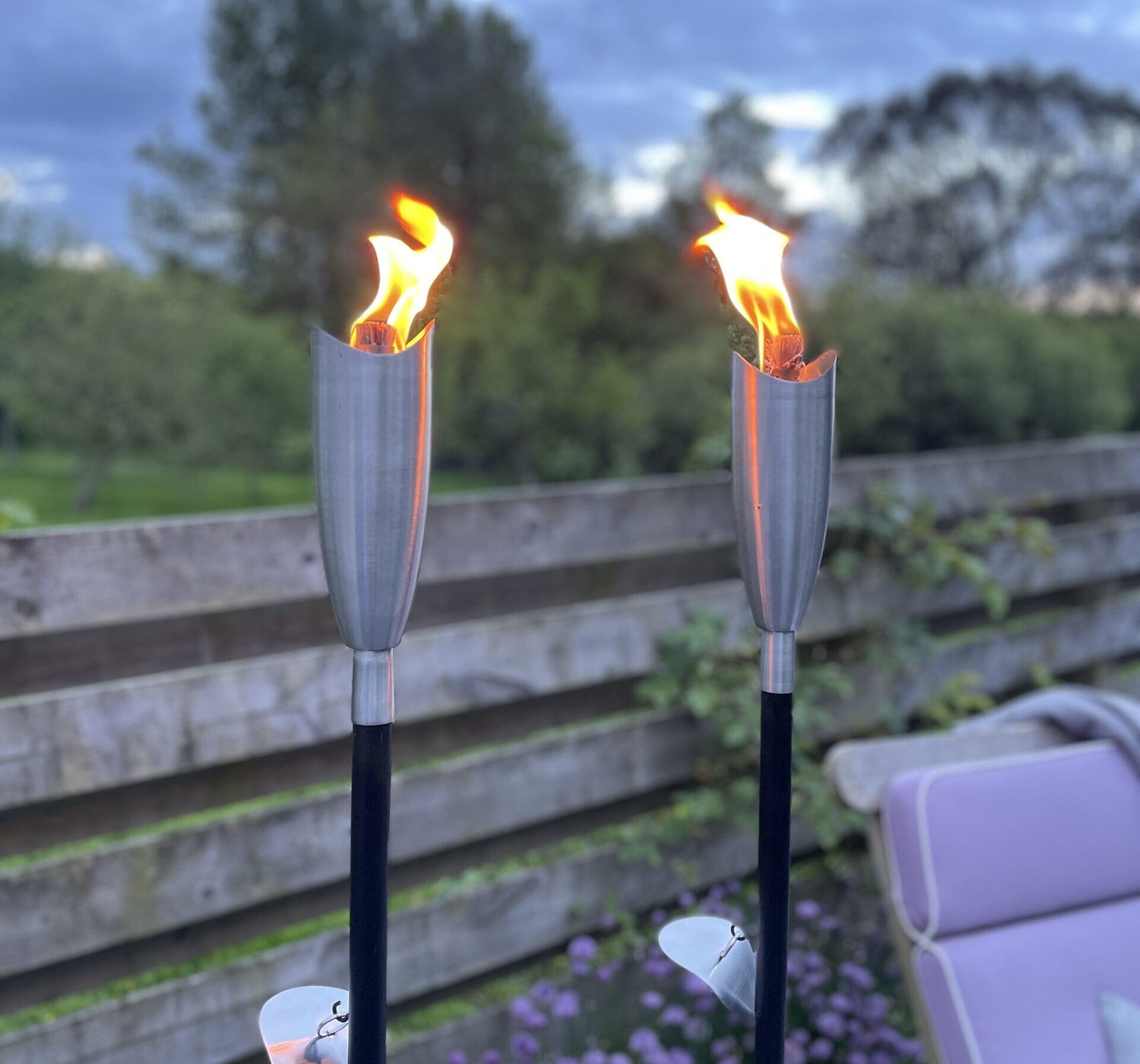 Silver oil torches in the evening, in the garden in front of flowers and chair.